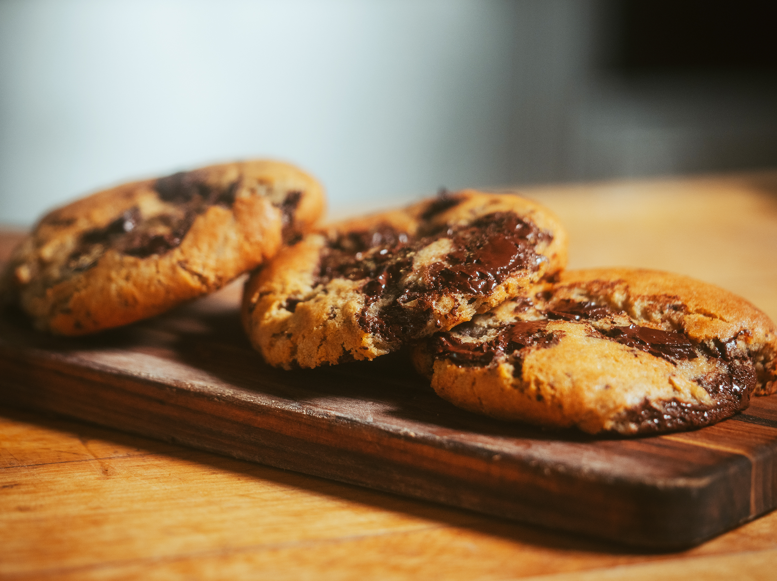 Three warm, freshly baked chocolate chip cookies from True Food Kitchen, placed on a wooden board, showcasing their gooey, melted chocolate chunks and golden-brown exterior.