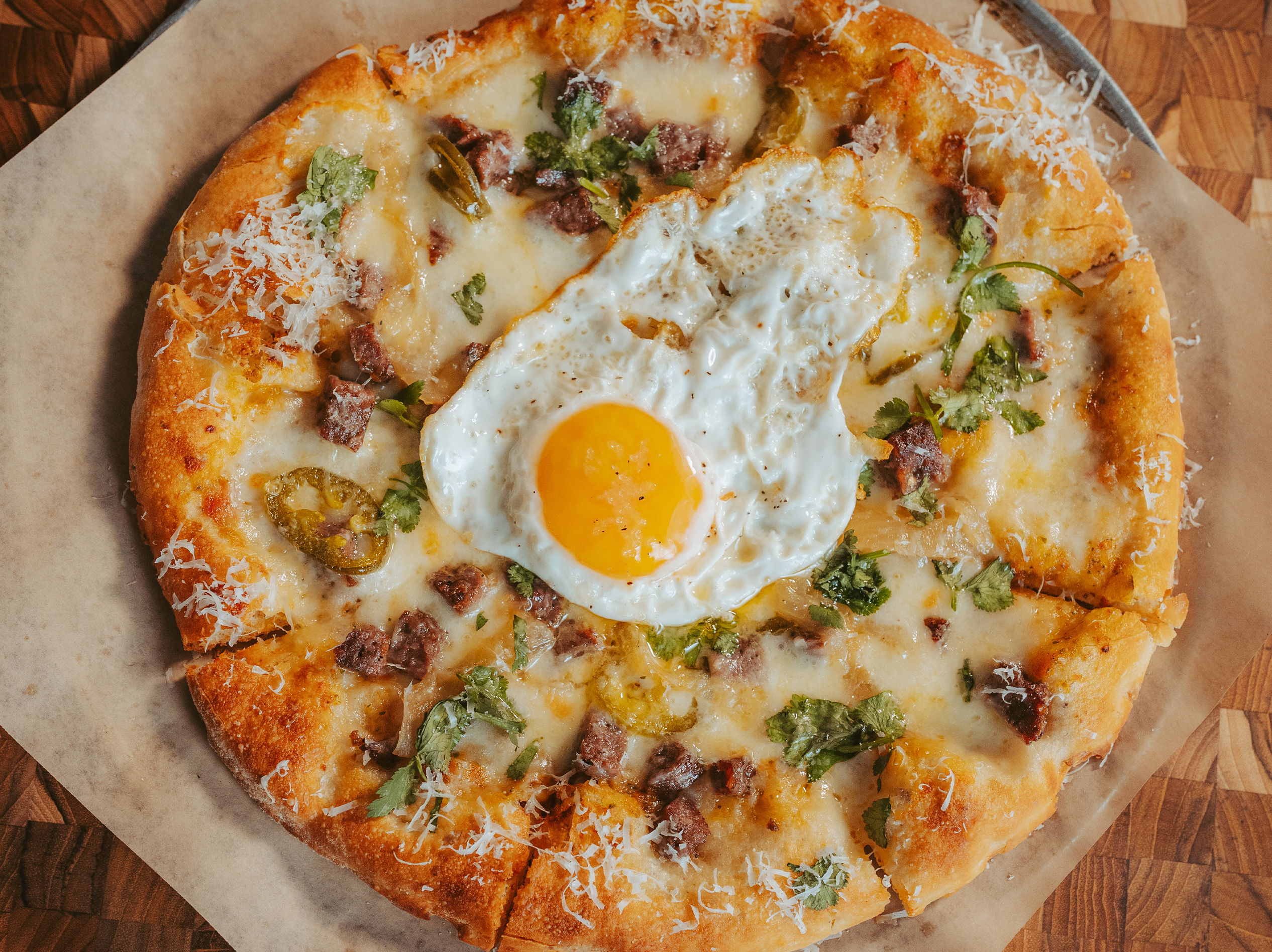 True Food Kitchen's sourdough pizza topped with melted cheese, sausage pieces, jalapeno slices, and a sunny-side-up egg, served on parchment paper.