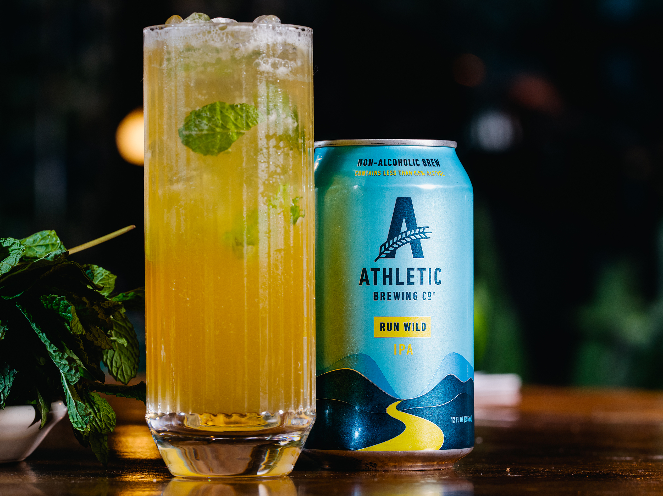 A tall glass of iced, non-alcoholic cocktail garnished with mint leaves, placed next to a can of Athletic Brewing Co.'s Run Wild IPA, on a wooden surface with a dark background.