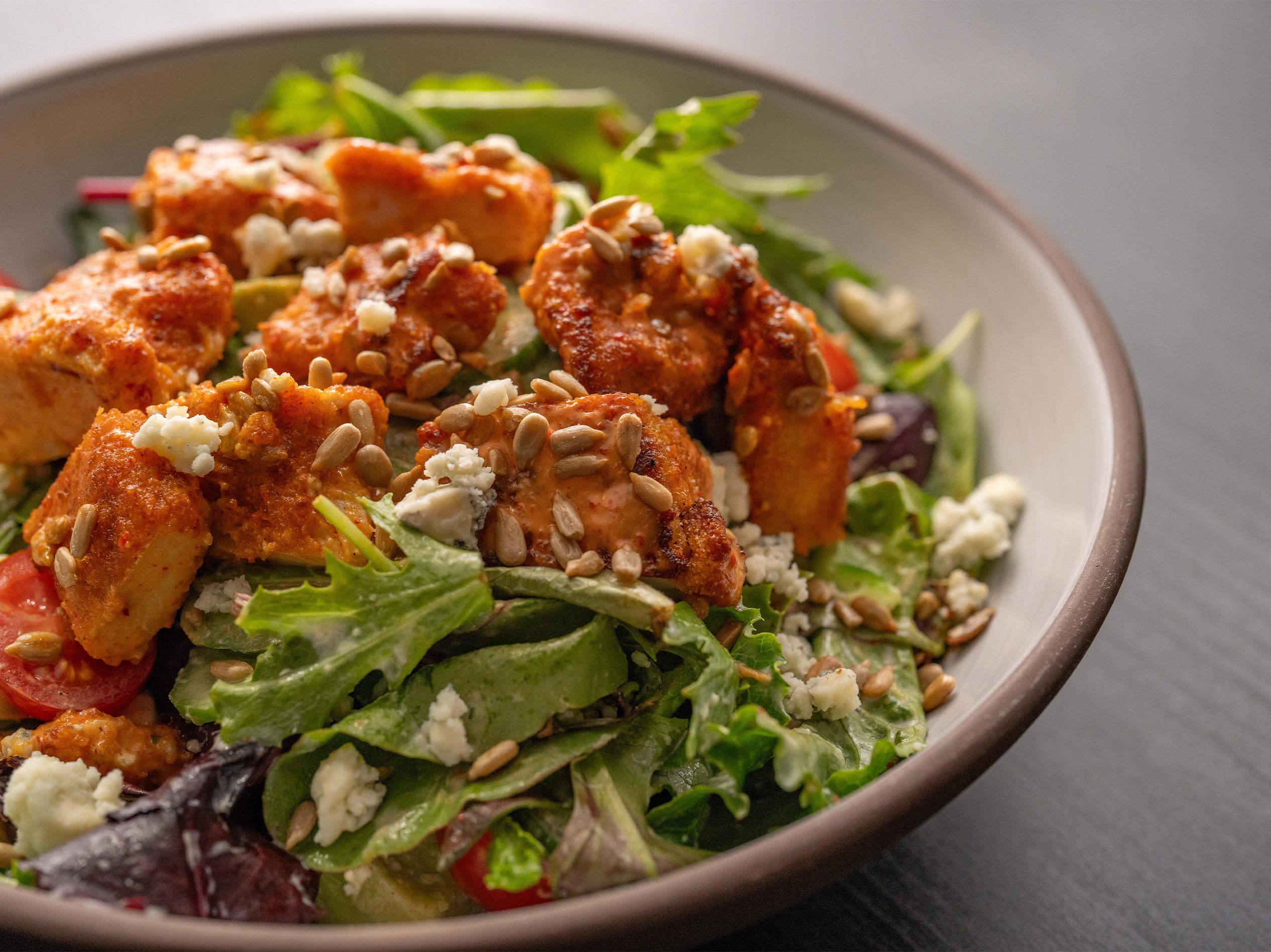 A bowl of Buffalo Chicken Ranch Salad featuring organic mixed greens, Point Reyes blue cheese, avocado, cherry tomatoes, cucumber, and organic sunflower seeds, set against an orange background.