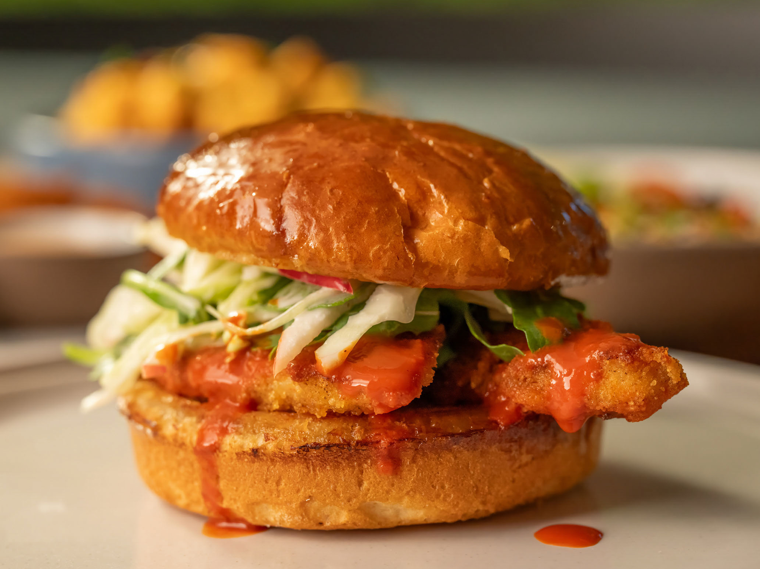 The Spicy Korean Chicken Sandwich from True Food Kitchen, featuring crispy chicken in a brioche bun with house-made pickles, Korean BBQ sauce, and kale slaw, set against an orange background.