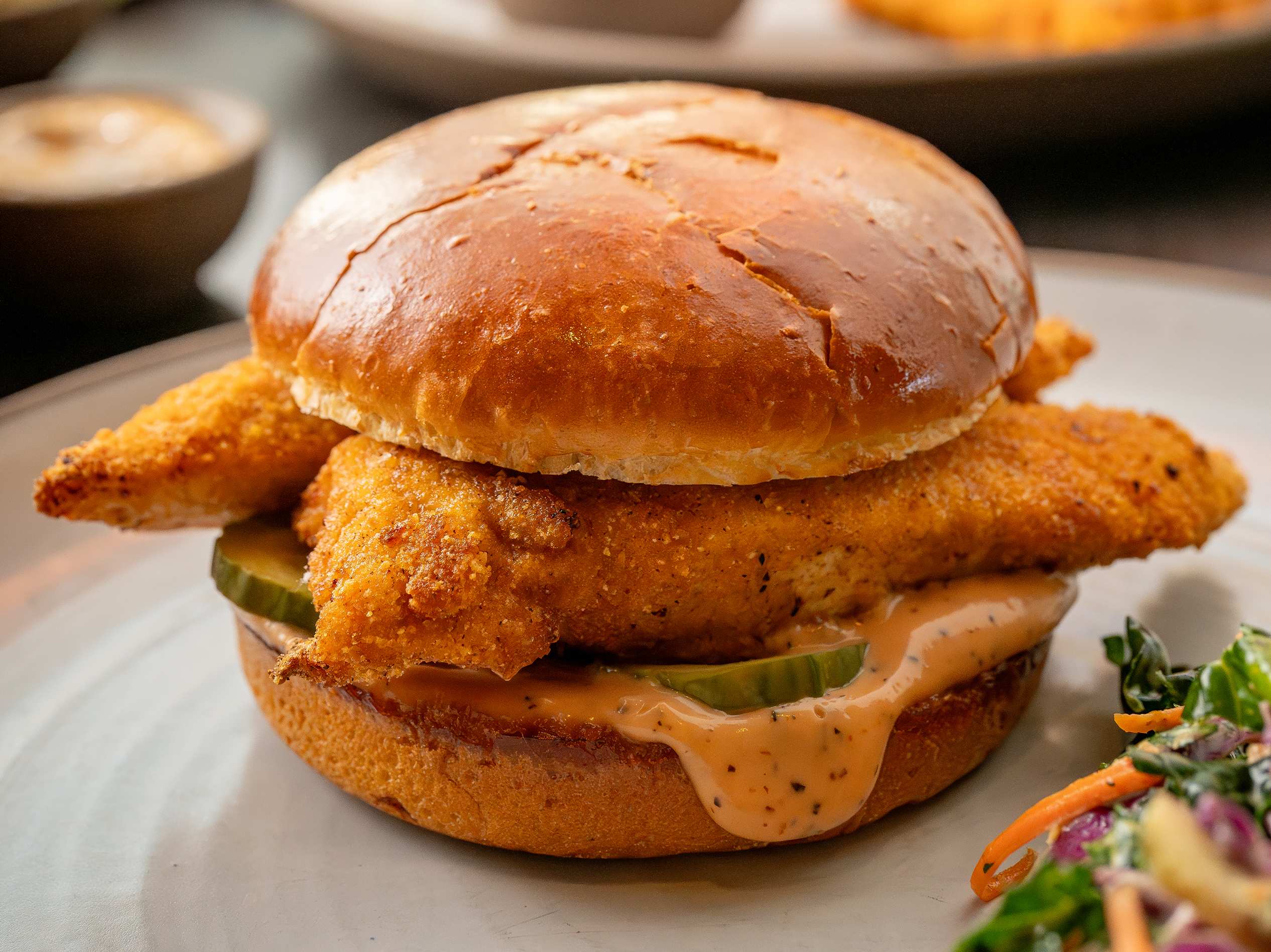 The Original Chicken Sandwich from True Food Kitchen, showing a hand holding a brioche bun filled with crispy chicken, house-made pickles, and awesome sauce, served with a choice of kale salad or simple green salad, set against an orange background.