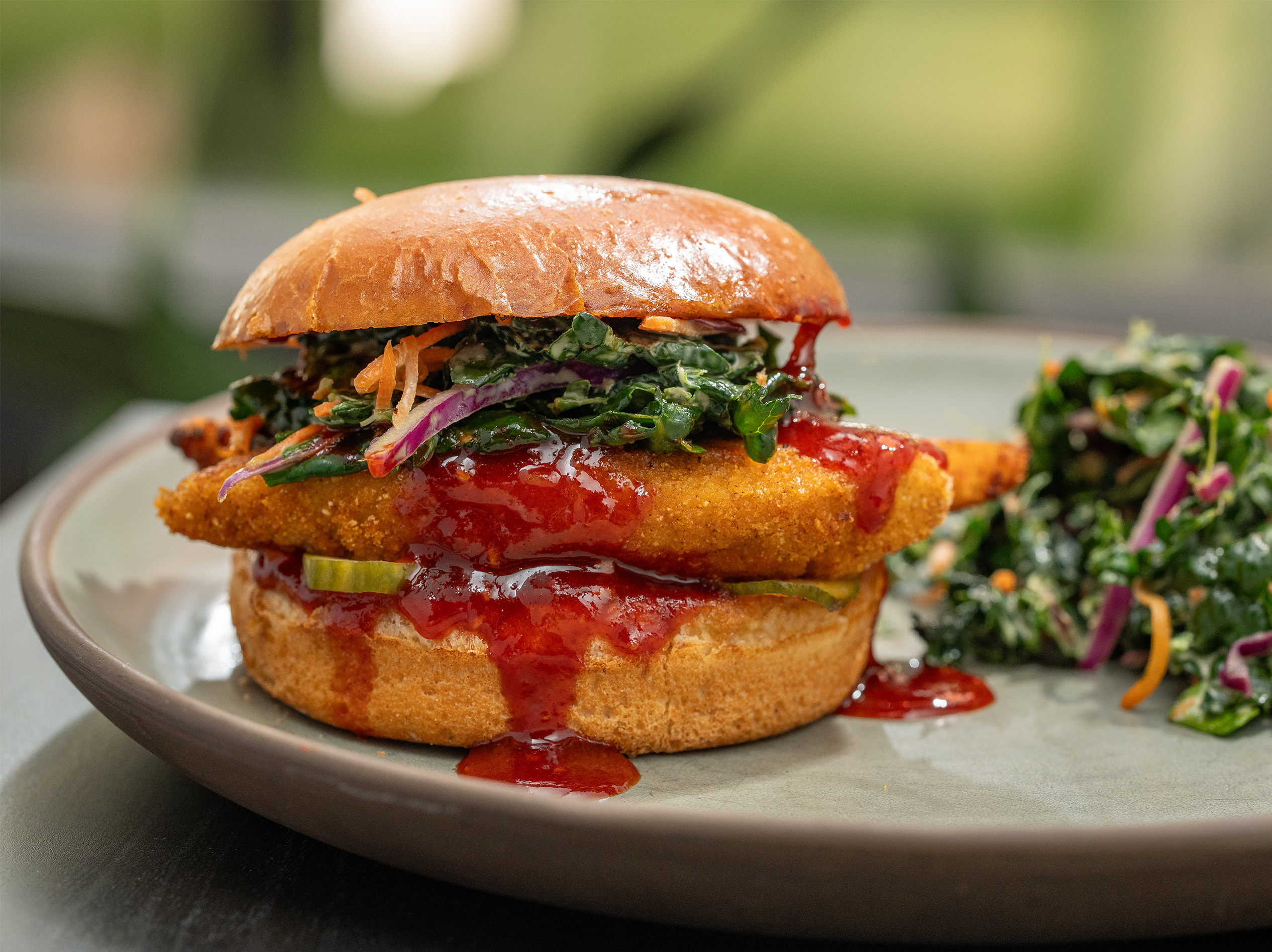 The Spicy Korean Chicken Sandwich from True Food Kitchen, featuring crispy chicken in a brioche bun with house-made pickles, Korean BBQ sauce, and kale slaw, set against an orange background.