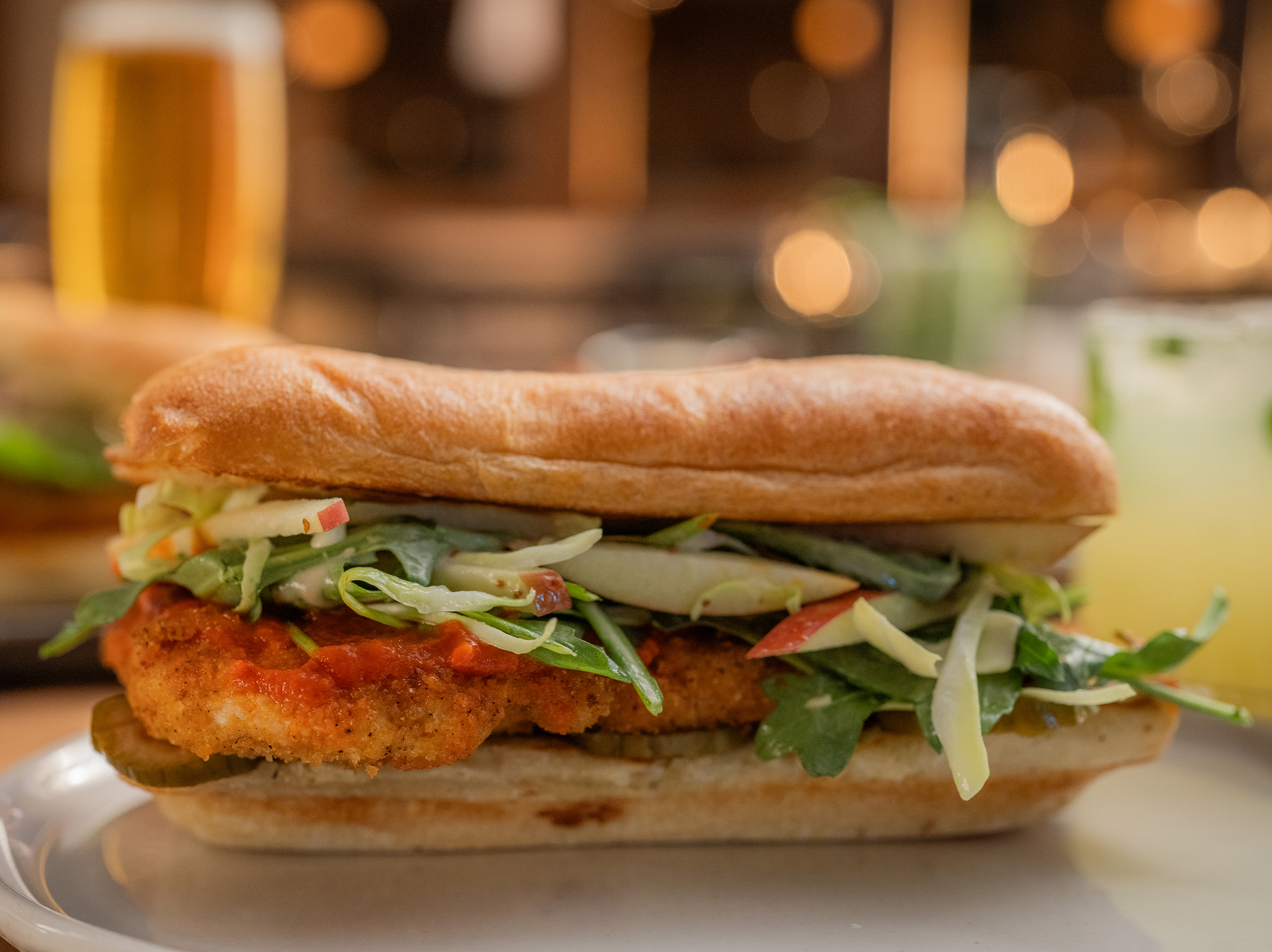 The Spicy Korean Chicken Sandwich from True Food Kitchen, featuring crispy chicken in a brioche bun with house-made pickles, Korean BBQ sauce, and kale slaw, set against an orange background.